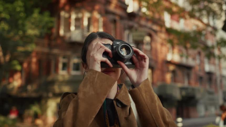 A young woman stands on a picturesque city street, raising her vintage camera to take a photograph in the warm afternoon light