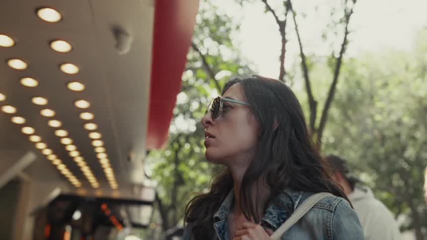 A stylish young woman in sunglasses stands on a city street and curiously looks up at a brightly lit building facade