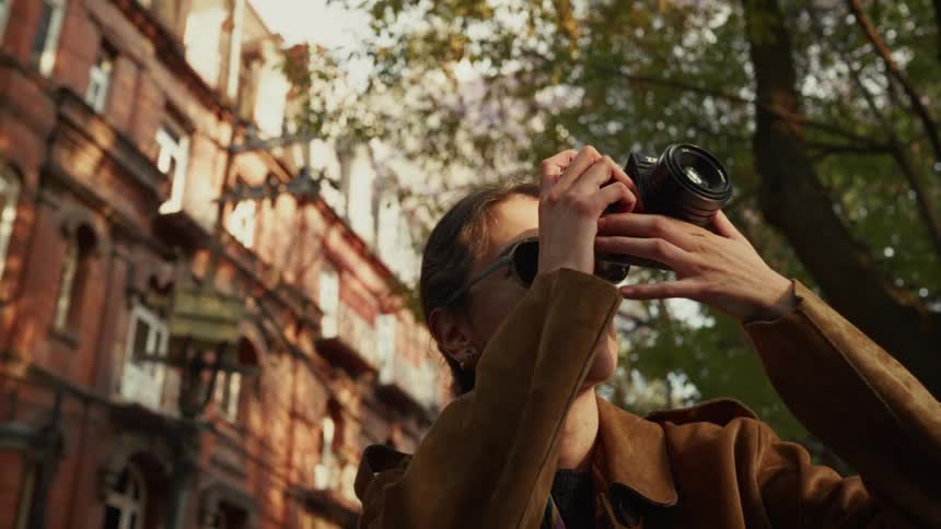 A low-angle view of a stylish young woman in sunglasses taking a photograph with her film camera on a city street