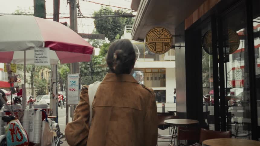 A cinematic slow-motion shot follows an older woman with gray hair walking down a city street in Mexico City