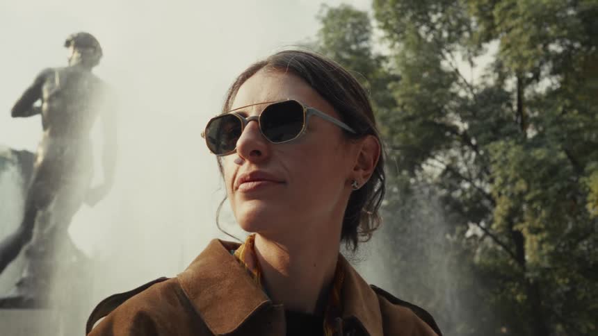 Low angle shot of a stylish woman in sunglasses and a brown coat looking on in front of a misty fountain and a large statue