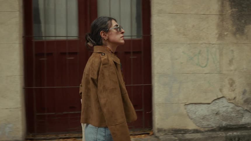 A stylish woman wearing a brown jacket and sunglasses walks along a weathered city building in slow motion