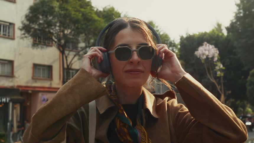 A fashionable woman puts on headphones while standing on a sunlit city street