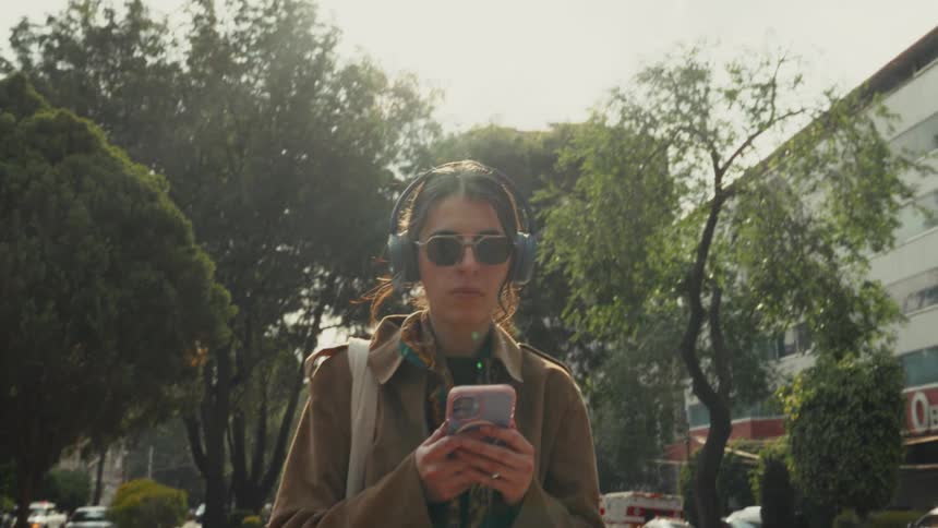 A stylish young woman wearing headphones and sunglasses walks towards the camera in slow motion through a sunny city park while looking at her smartphone