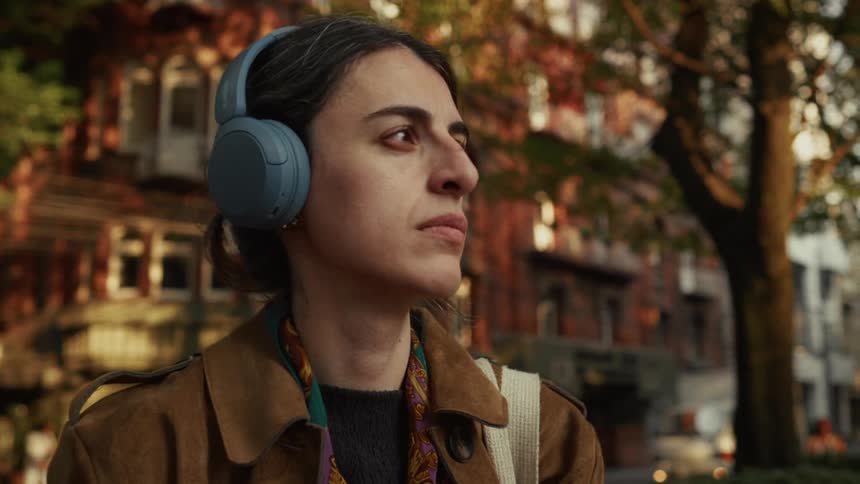 A thoughtful woman with headphones lowers her phone after taking a picture on a tree-lined city street at golden hour