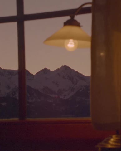 A cozy lamp illuminates a warm room with a view of snow-capped mountains at dusk through the window
