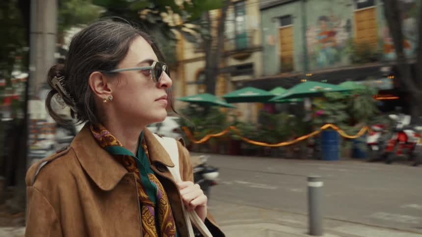 A stylish woman wearing sunglasses and a brown coat walks in slow motion along a colorful city street
