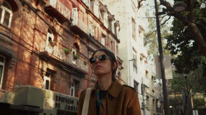 A stylish woman in sunglasses walks along a sunlit city street past historic brick buildings in slow motion