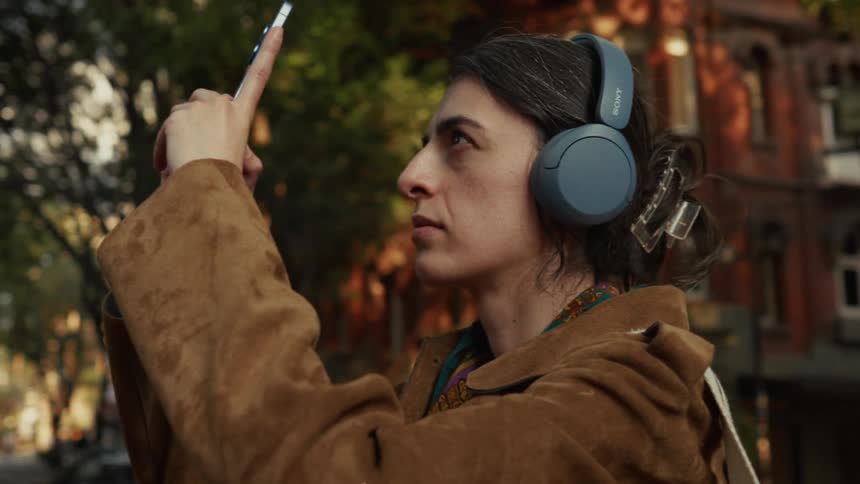 From a low angle, a woman with headphones holds her phone up to take a picture of the city buildings at golden hour
