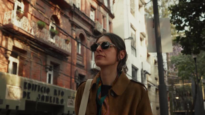 A stylish woman wearing sunglasses and a coat confidently walks through a historic city street in the warm afternoon sun