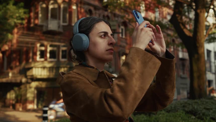 Cinematic slow-motion of a woman with headphones taking a video of a historic brick building with her smartphone in the city
