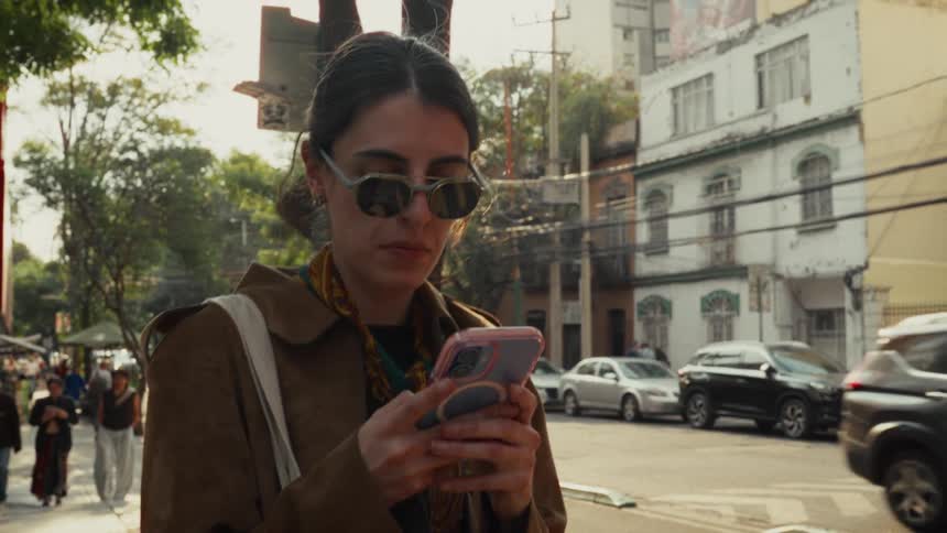 A fashionable young woman wearing sunglasses stands on a bustling city sidewalk, absorbed in her smartphone as traffic passes by