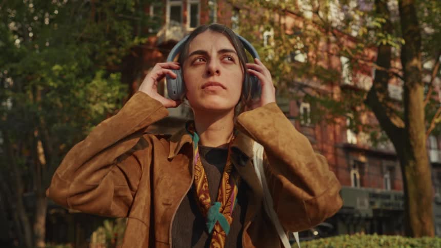 A young woman with a thoughtful expression puts on her headphones and walks away on a sunny city street