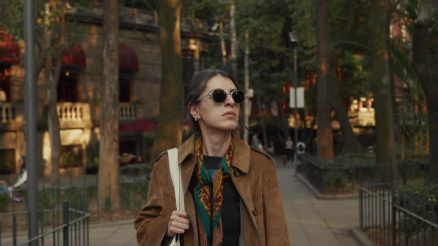 A fashionable young woman wearing a brown coat and sunglasses walks thoughtfully through a sunlit city park