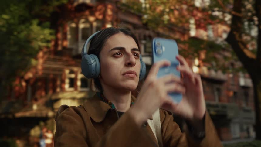 A young woman wearing blue headphones takes a photo with her smartphone on a sun-dappled city street in slow motion