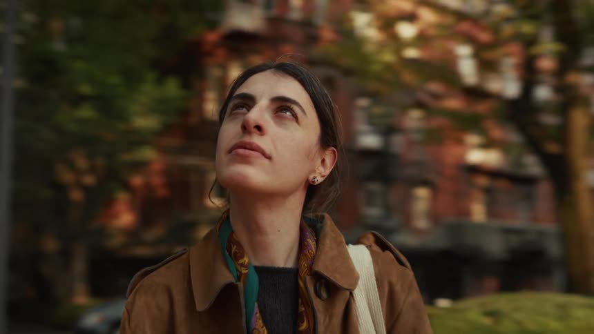 A serene woman in a brown coat looks up in wonder while standing in a lush city park as the camera orbits her