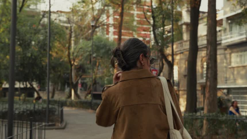 A slow-motion, over-the-shoulder shot of a woman with graying hair walking through a city park in the afternoon