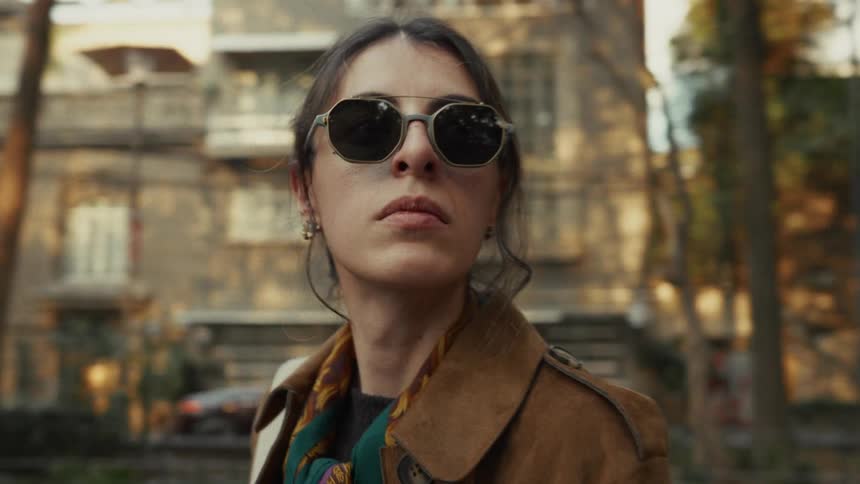 A stylish woman in sunglasses and a trench coat walks with purpose along a city park path as the camera circles to her face