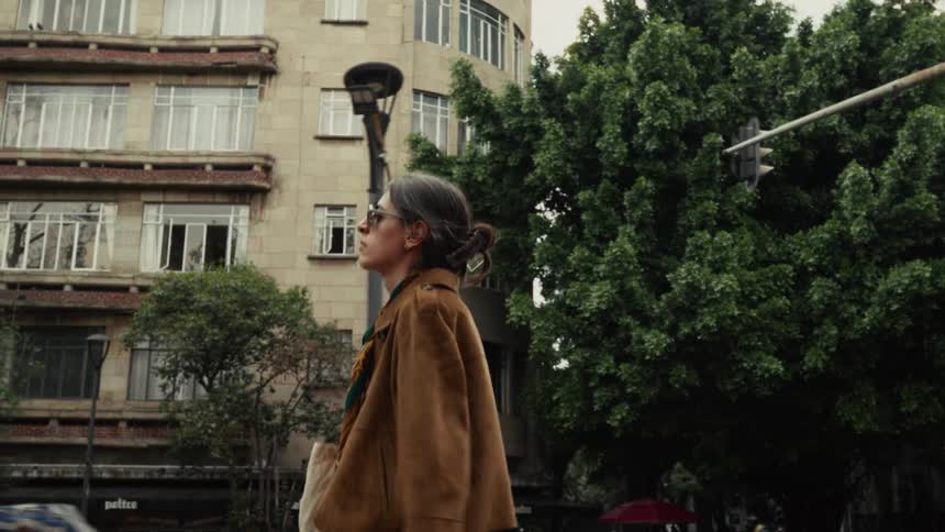 A stylish woman in sunglasses and a brown jacket confidently walks down a city street in slow motion