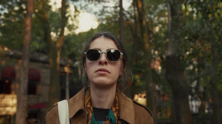 Cinematic low-angle portrait of a stylish woman in sunglasses standing thoughtfully in a park at golden hour