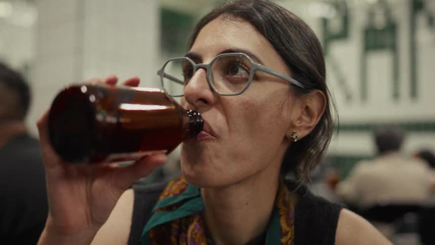 Close-up of a contemplative woman in glasses slowly drinking from a bottle in a bustling diner