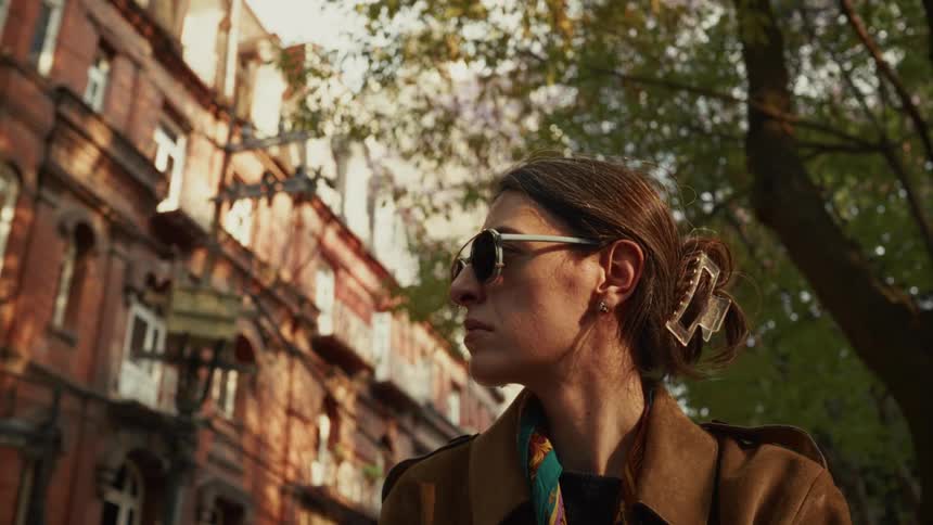 A low-angle shot captures a stylish woman in sunglasses looking around a city street bathed in the warm glow of the setting sun