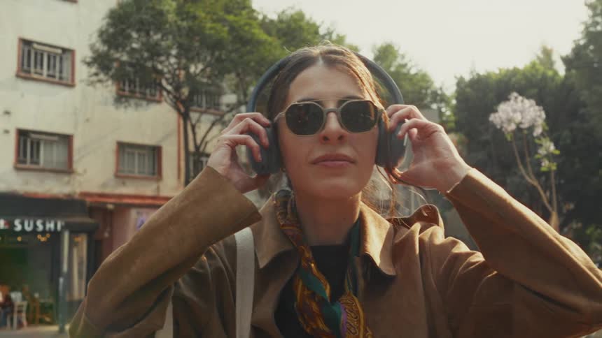 A stylish woman in sunglasses puts on headphones while standing on a sunny city street in slow motion