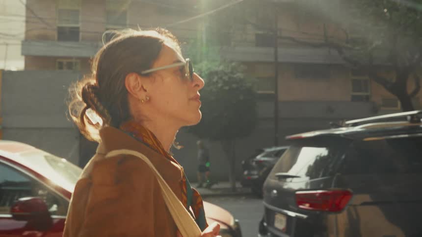 Side profile of a stylish woman in sunglasses walking down a sunny city street in slow motion