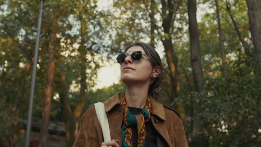 A stylish woman in sunglasses and a brown coat looks up thoughtfully while standing in a sun-dappled park
