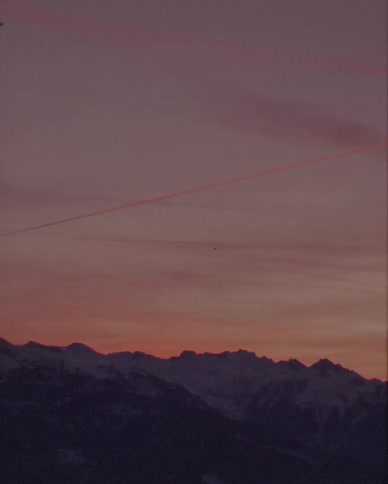 A vintage shot of airplane contrails streaking across a colorful sunset sky over a snow-covered mountain range