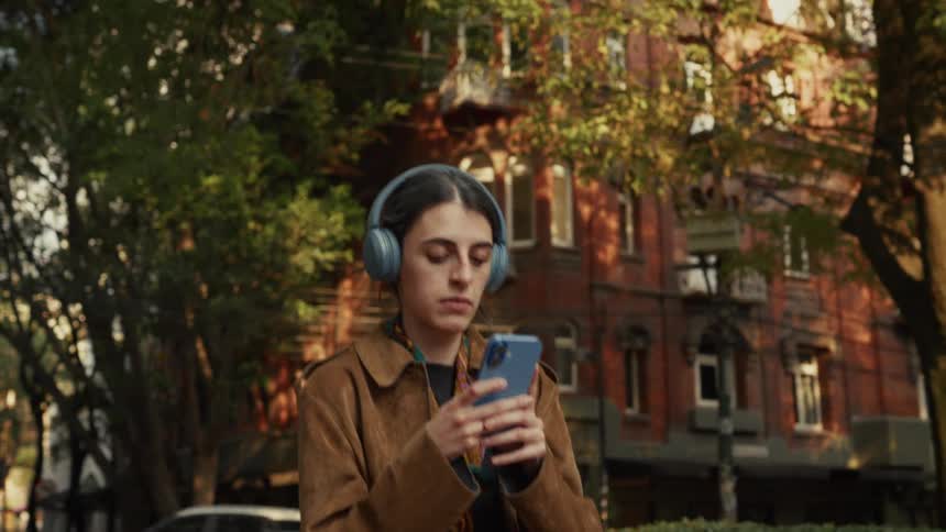 A young woman wearing a brown coat and headphones scrolls through her smartphone while walking down a sunny, tree-lined city street