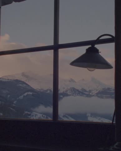 A silhouetted lamp hangs in a window framing a breathtaking view of snow-covered mountains shrouded in clouds