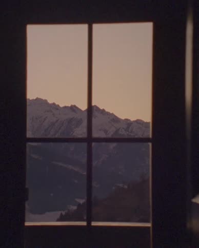 A warm light leak transitions to a calm, picturesque view of snow-capped mountains through a window at sunrise