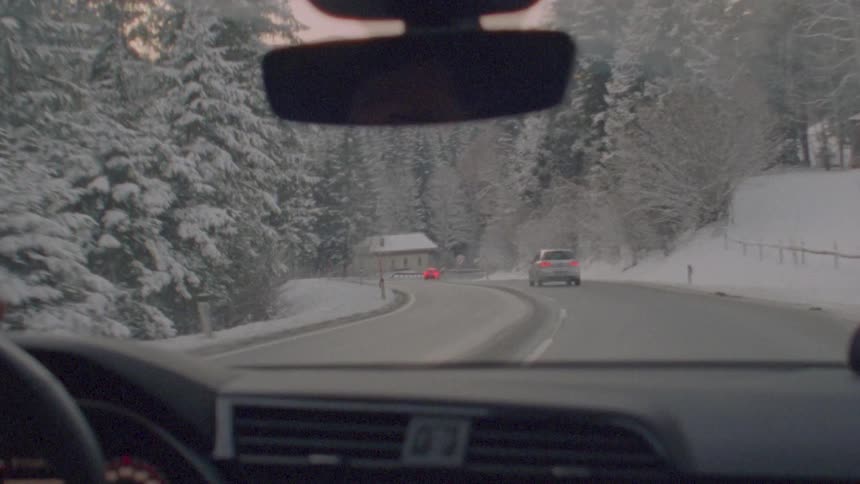 A point-of-view shot from a car driving on a winding road through a snowy mountain forest at dusk