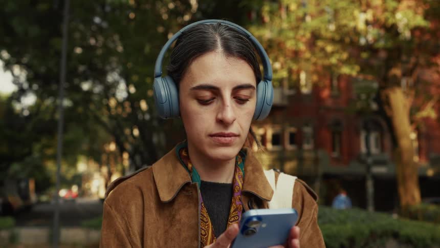 A focused young woman wearing headphones looks at her smartphone while standing in a sunlit city park before walking away