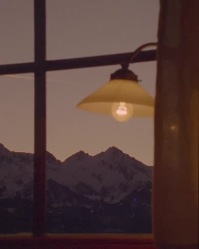 A cozy interior view of snow-capped mountains at sunset through a window, with a warm lamp glowing in the foreground