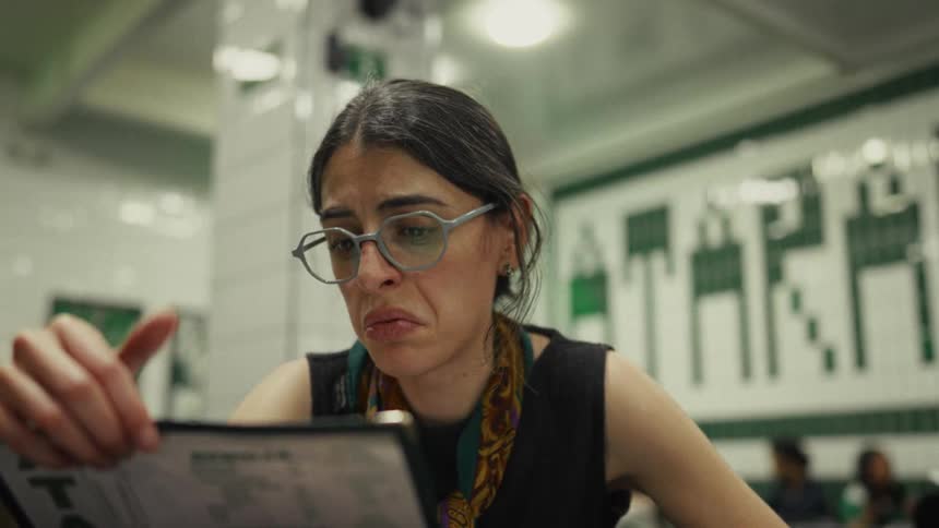 A stylish woman with glasses intently reads a menu in a brightly lit diner before pointing out her order