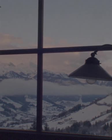 A slow pan across a window with a silhouetted lamp reveals a vast, snowy mountain valley at dusk