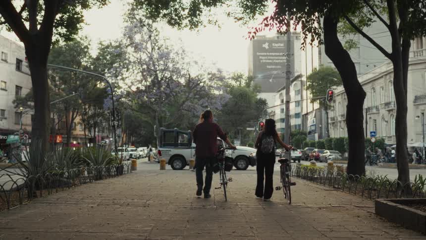 A man and a woman walk down a tree-lined city sidewalk with their bicycles towards a busy street