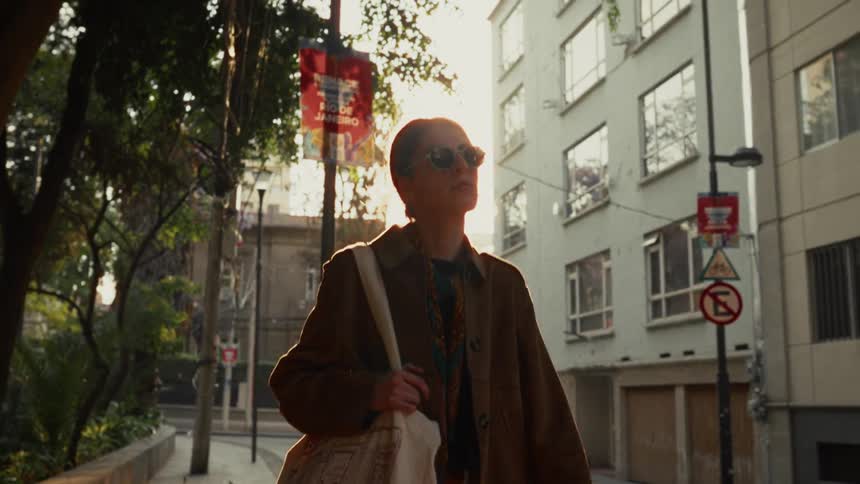 A young woman wearing sunglasses walks along a city sidewalk, beautifully backlit by the golden hour sun