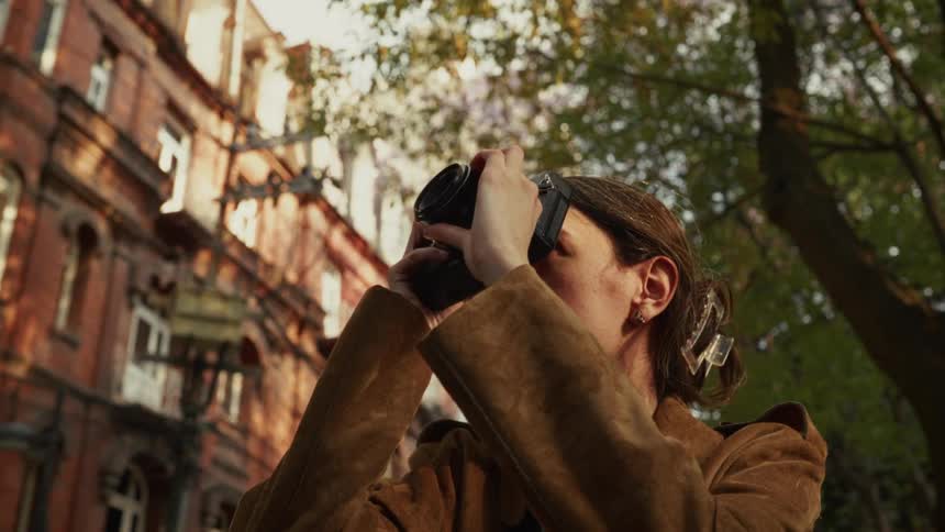A low-angle shot of a woman in a brown coat raising her camera to photograph a historic brick building during golden hour
