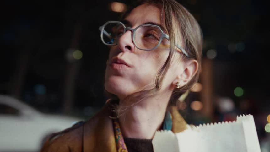 Slow-motion close-up of a young woman in glasses savoring a churro on a windy city night