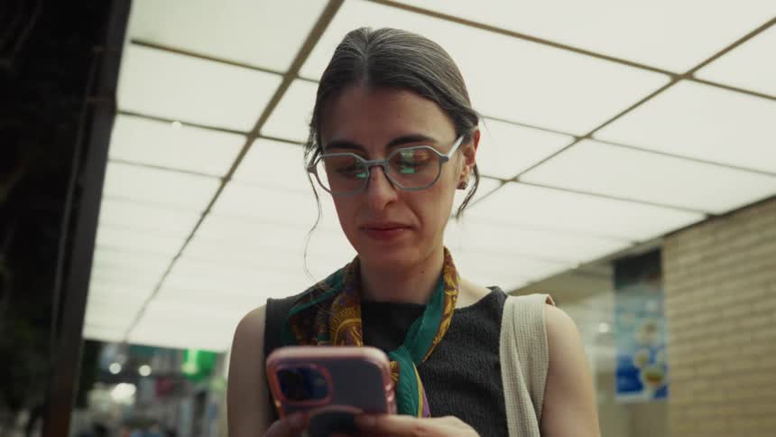 A woman with glasses and a stylish scarf uses her smartphone under a brightly lit overhang on a city street in the evening