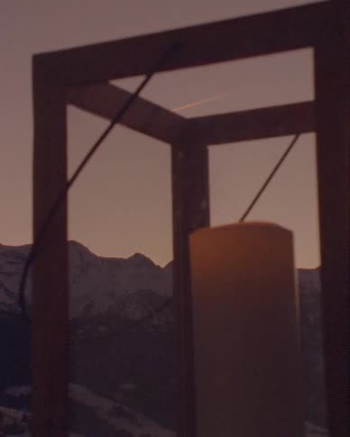 A tranquil close-up of a candle in a wooden lantern with snowy mountains visible in the background during a warm sunset