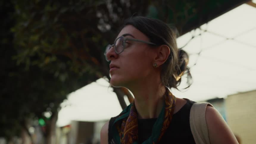 A young woman with glasses looks up from her phone to scan her surroundings while waiting on a city street