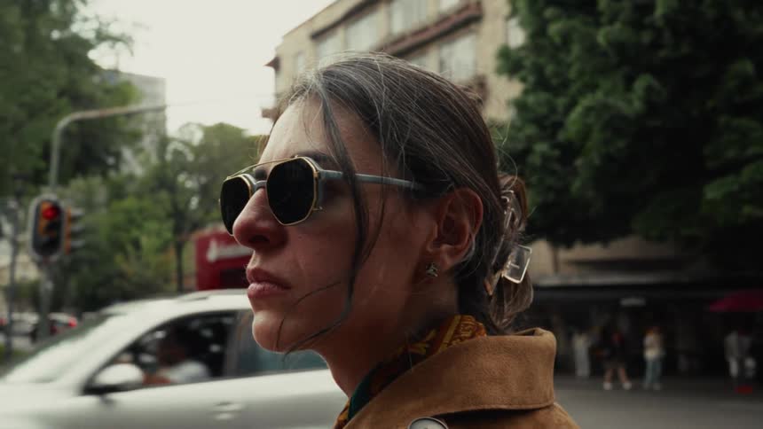 A fashionable woman in a brown coat and sunglasses stands contemplatively on a city street