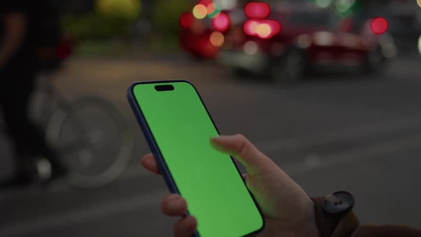 A close-up of a person's hand scrolling on a smartphone with a green screen on a bustling city street at dusk