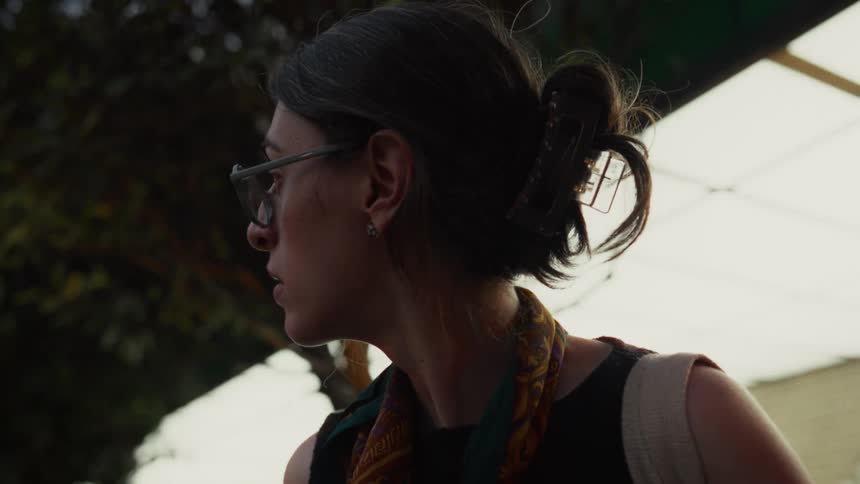 A pensive woman with glasses waits at a public transport stop, looking around with a concerned expression