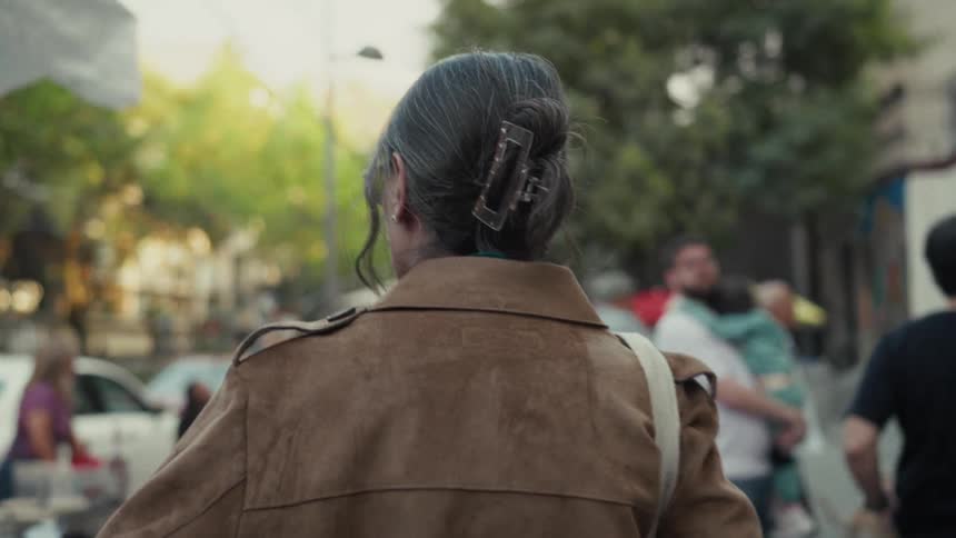A woman with her hair in a claw clip is seen from behind walking through a bustling outdoor city market in slow motion