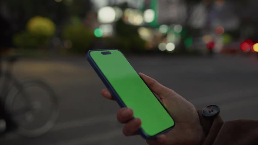 Over-the-shoulder shot of a person's hand holding a smartphone with a green screen on a blurry city street at night